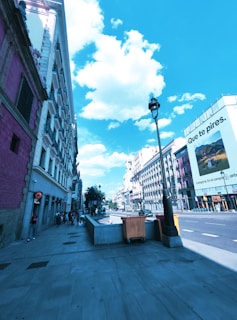 A city street with people walking on the sidewalk next to multistory buildings. The sky is bright blue with clouds, and there is a large billboard featuring Spanish text. A streetlamp and some trash bins are visible, and the scene has modern urban architecture.
