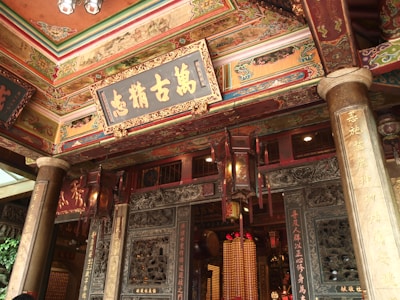 An intricately decorated traditional structure with detailed gold and colorful designs. The ceilings are adorned with ornate patterns and there are large wooden lanterns hanging. Chinese characters are inscribed on wooden plaques and pillars, adding to the cultural and historical ambiance.