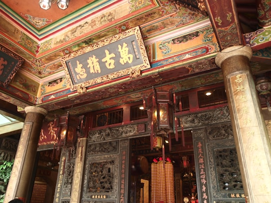 An intricately decorated traditional structure with detailed gold and colorful designs. The ceilings are adorned with ornate patterns and there are large wooden lanterns hanging. Chinese characters are inscribed on wooden plaques and pillars, adding to the cultural and historical ambiance.