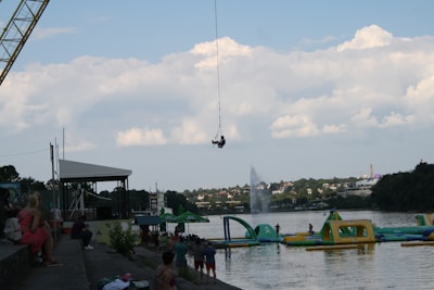 A bungee jump attraction is set up by a river, with a person mid-jump suspended on a cable. The scene includes an inflatable water park in the river with people interacting with the structures. On the left, spectators sit on stone steps watching the activities. Dense clouds sit in a blue sky above a tree-lined embankment across the river with buildings visible in the distance.