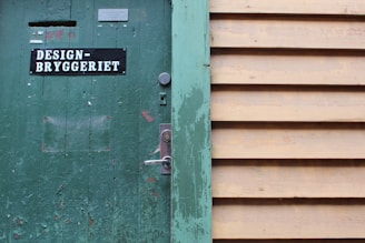 A weathered green wooden door with signs of peeling paint, featuring a sign with the text 'DESIGN-BRYGGERIET'. The door has a silver lock and handle. Adjacent to the door is a wall made of horizontal beige wooden slats.