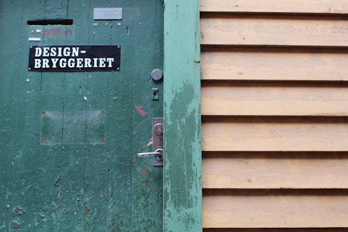 A weathered green wooden door with signs of peeling paint, featuring a sign with the text 'DESIGN-BRYGGERIET'. The door has a silver lock and handle. Adjacent to the door is a wall made of horizontal beige wooden slats.