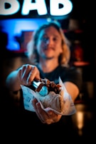 A person is holding a wrapped gyro or pita sandwich filled with meat, using a pair of tongs. The background features a brightly lit sign with the letters 'DAB'. The surroundings are dimly lit, creating a focused lighting effect on the food.