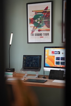 A modern workspace featuring a desktop computer, laptop on a stand, and a tablet on a wooden desk. A framed art poster titled 'The Grand Tour' is displayed on the wall. A sleek desk lamp provides additional lighting. Books and a small speaker are also present, contributing to the aesthetic setup.