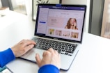 A person wearing a blue sleeve types on a MacBook Pro. The laptop screen displays a website editing interface with beauty products and a smiling woman applying makeup.