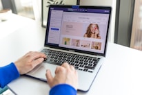 A person wearing a blue sleeve types on a MacBook Pro. The laptop screen displays a website editing interface with beauty products and a smiling woman applying makeup.
