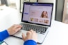 A person wearing a blue sleeve types on a MacBook Pro. The laptop screen displays a website editing interface with beauty products and a smiling woman applying makeup.