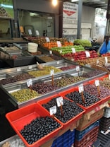 A market stall displaying a variety of fresh fruits and olives. The stall features rows of colorful produce, including green, yellow, and red fruits such as apples and oranges. Several bins contain different types of olives, each labeled with a price. The background has market signs and a vendor in the corner.