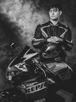 A person in a motorcycle racing suit stands confidently, resting hands on a helmet placed on a sport motorcycle. The bike features a Repsol Honda design. The setting is dramatic with a textured, cloudy backdrop.