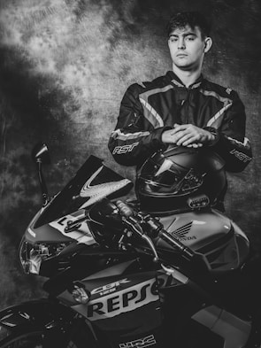 A person in a motorcycle racing suit stands confidently, resting hands on a helmet placed on a sport motorcycle. The bike features a Repsol Honda design. The setting is dramatic with a textured, cloudy backdrop.