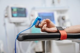 A person's hand resting on a chair arm while wearing a blue clip device connected to a cable. Medical monitors with blurred displays are in the background, indicating a hospital or clinical setting. A red strap is visible on the chair arm, suggesting stability or restraint for a procedure.