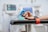 A person's hand resting on a chair arm while wearing a blue clip device connected to a cable. Medical monitors with blurred displays are in the background, indicating a hospital or clinical setting. A red strap is visible on the chair arm, suggesting stability or restraint for a procedure.
