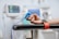 A person's hand resting on a chair arm while wearing a blue clip device connected to a cable. Medical monitors with blurred displays are in the background, indicating a hospital or clinical setting. A red strap is visible on the chair arm, suggesting stability or restraint for a procedure.