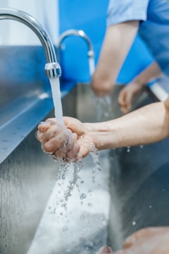 Hands are under running water from a faucet, with droplets splashing in a stainless steel sink. The setting appears clean and sanitary, with a focused action on handwashing.