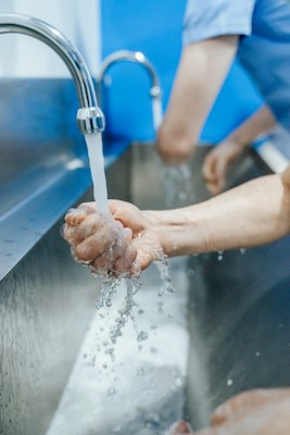 Hands are under running water from a faucet, with droplets splashing in a stainless steel sink. The setting appears clean and sanitary, with a focused action on handwashing.