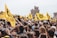 A large crowd of people gathered outdoors, holding and waving yellow flags with a logo and text on them. The setting appears to be near a historic stone tower and other architectural structures. The atmosphere is lively and festive, with people looking engaged and cheerful.