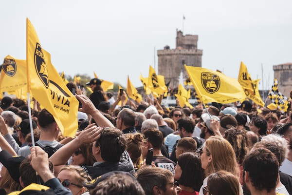 A large crowd of people gathered outdoors, holding and waving yellow flags with a logo and text on them. The setting appears to be near a historic stone tower and other architectural structures. The atmosphere is lively and festive, with people looking engaged and cheerful.