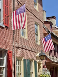 A historic brick building with two Betsy Ross flags displayed outside. The building features red, green, and cream-colored window shutters and an old-fashioned street lamp with hanging plants in the foreground.