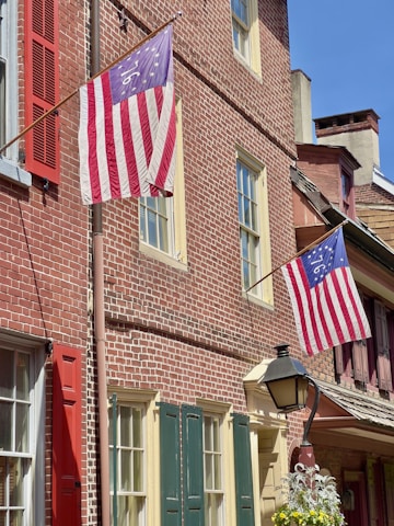 A historic brick building with two Betsy Ross flags displayed outside. The building features red, green, and cream-colored window shutters and an old-fashioned street lamp with hanging plants in the foreground.