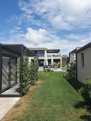 A modern house with multiple sections, surrounded by greenery and a well-maintained lawn. The architecture features clean lines and large glass windows. There are white umbrellas shading a seating area on a patio.