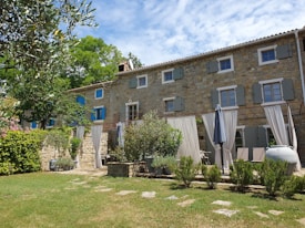 A rustic stone house with several windows featuring closed shutters, enveloped by greenery. The courtyard has a grassy lawn, trimmed hedges, and potted plants. A series of white outdoor curtains hang along the patio area, next to various seating arrangements and a closed umbrella. The sky is partly cloudy, providing a serene and inviting atmosphere.