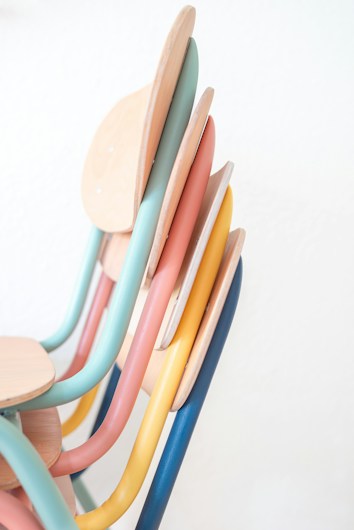 Four stacked chairs with multicolored metal frames and wooden seats. The chairs feature pastel shades of blue, pink, yellow, and green, highlighting a modern and playful design.