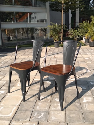 Two metal chairs with wooden seats are placed on a tiled patio. Potted plants and a modern building with large windows are visible in the background.
