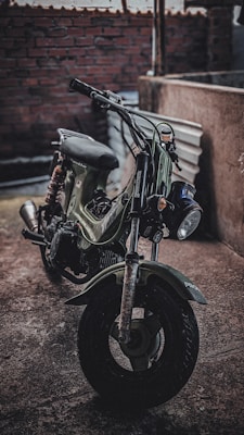 A vintage motorcycle with a rugged, weathered appearance is parked in a dimly lit, rough-textured urban environment. The motorcycle has an olive-green body and is positioned at an angle, showcasing its handlebars and front tire. The background includes brickwork and concrete surfaces, adding to the industrial feel.
