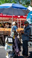 A street market scene with various vendors under red tents displaying a large array of fruits, including peaches. A person wearing a blue checkered shirt and a cap is standing beside a cart that has donation signs and various items attached to it. There is a large blue umbrella providing shade.