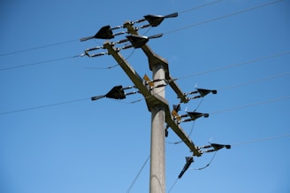 A tall concrete utility pole is equipped with several electrical insulators and wires against a clear blue sky. The pole is used for the distribution of electrical power and has various metal components attached to it.