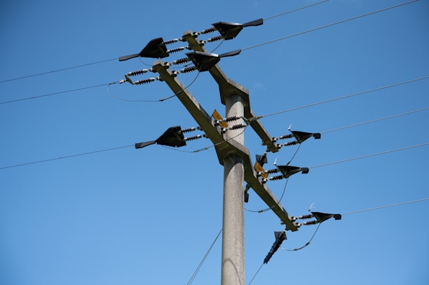 A tall concrete utility pole is equipped with several electrical insulators and wires against a clear blue sky. The pole is used for the distribution of electrical power and has various metal components attached to it.