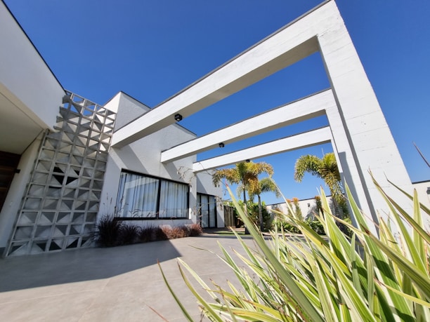 A modern architectural building with geometric design elements, large windows, and visible structural beams. There are palm trees and various plants around the entrance area, with a clear blue sky overhead.