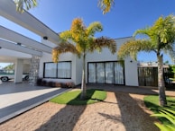 A modern house with a white facade and large windows, bordered by palm trees. The front features a mix of gravel and grass landscaping, complemented by a clear blue sky and a covered carport area with a vintage car visible in the corner.