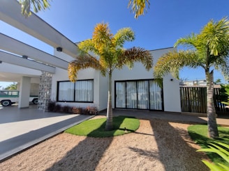 A modern house with a white facade and large windows, bordered by palm trees. The front features a mix of gravel and grass landscaping, complemented by a clear blue sky and a covered carport area with a vintage car visible in the corner.