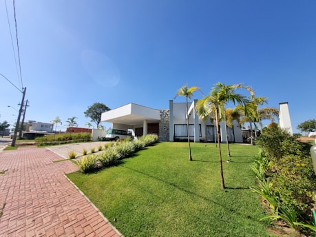 A modern, white architectural building with large windows, surrounded by a well-maintained green lawn and several palm trees. The sky is clear and blue, indicating a sunny day. A paved brick pathway leads up to the building, and a vintage van is parked under a shaded area.