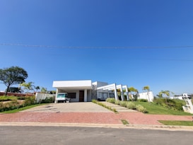 A modern, single-story house with a flat roof and a minimalist design. The structure includes a carport with a white vehicle parked underneath, large glass windows, and a landscaped front yard with palm trees and bushes. The sky is clear and blue, adding a sense of openness.