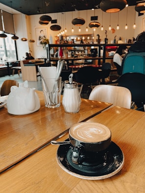 A cozy cafe setting featuring a latte with latte art in a black cup and saucer on a wooden table. The background showcases a warm and inviting cafe ambiance with hanging pendant lights, framed pictures on the wall, and various patrons seated at tables.