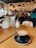 A cozy cafe setting featuring a latte with latte art in a black cup and saucer on a wooden table. The background showcases a warm and inviting cafe ambiance with hanging pendant lights, framed pictures on the wall, and various patrons seated at tables.