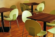 A room contains several green chairs and dark wooden tables arranged on a polished wooden floor. The setting is minimalistic, potentially part of a cafe or small dining area. The chairs have a modern, rounded design with metal legs, and the tables are square with sturdy bases.