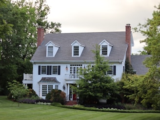 A large, two-story house with white siding and dark shutters is surrounded by lush greenery. The building features a gabled roof with dormer windows and brick chimneys on either side. The front yard has a well-maintained lawn and landscaped bushes.