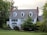 A large, two-story house with white siding and dark shutters is surrounded by lush greenery. The building features a gabled roof with dormer windows and brick chimneys on either side. The front yard has a well-maintained lawn and landscaped bushes.
