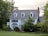 A large, two-story house with white siding and dark shutters is surrounded by lush greenery. The building features a gabled roof with dormer windows and brick chimneys on either side. The front yard has a well-maintained lawn and landscaped bushes.