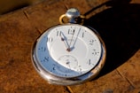 A vintage pocket watch with a white face and black numerals is placed on a textured, rusty surface. The watch appears to be made from a silver-colored metal and features classic hour, minute, and second hands.