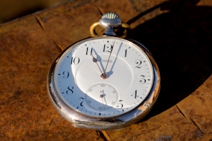 A vintage pocket watch with a white face and black numerals is placed on a textured, rusty surface. The watch appears to be made from a silver-colored metal and features classic hour, minute, and second hands.