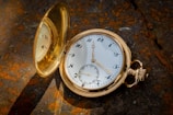A vintage gold pocket watch with an ornate exterior is open, revealing its classic white dial with black numerals and golden hands. The watch is set against a textured, rust-colored background, likely a stone surface with lots of natural light creating a warm and inviting atmosphere. The open lid reflects part of the dial and adds an element of depth to the scene.