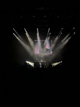 A DJ stands on a stage, illuminated by multiple beams of white light, creating a dynamic and energetic atmosphere. The audience is silhouetted in the foreground, with hands raised, suggesting a live music event.