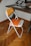 A bright orange folding chair is placed next to a wooden desk. The chair features a white frame with the word 'PANTONE 165' printed on the seat. The desk has a modern design with a keyboard, a raised monitor stand, and some books and other items in the background. The room has parquet flooring, which complements the warm tones of the chair.