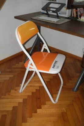 A bright orange folding chair is placed next to a wooden desk. The chair features a white frame with the word 'PANTONE 165' printed on the seat. The desk has a modern design with a keyboard, a raised monitor stand, and some books and other items in the background. The room has parquet flooring, which complements the warm tones of the chair.
