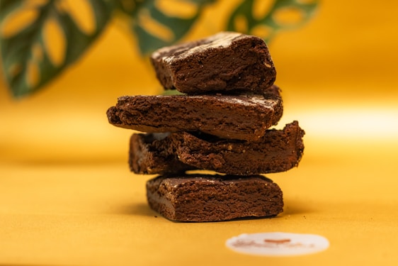 A stack of four rich, chocolate brownies placed against a warm, golden backdrop. The brownies appear dense and moist, with visible textures and a slightly glossy surface. In the background, part of a green leaf with a blurred effect adds contrast to the scene.