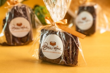 Individually wrapped chocolate brownies are neatly packaged in transparent plastic with an orange ribbon, placed against a warm yellow background. The brand label, featuring a stylized logo and text, is visible on the packaging.
